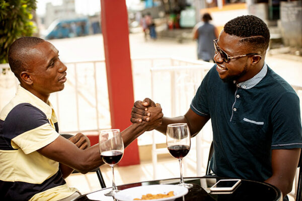 2 men in a cafe clasping hands in greeting, red wine on table